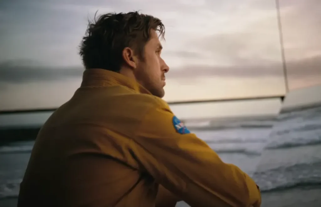 A still from Project Hail Mary. Ryland Grace is looking at screens that show the beach.